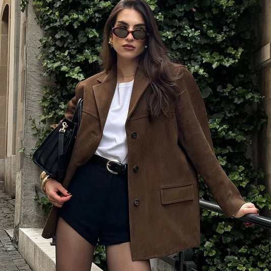 Woman in a brown coat and sunglasses standing against a green hedge.