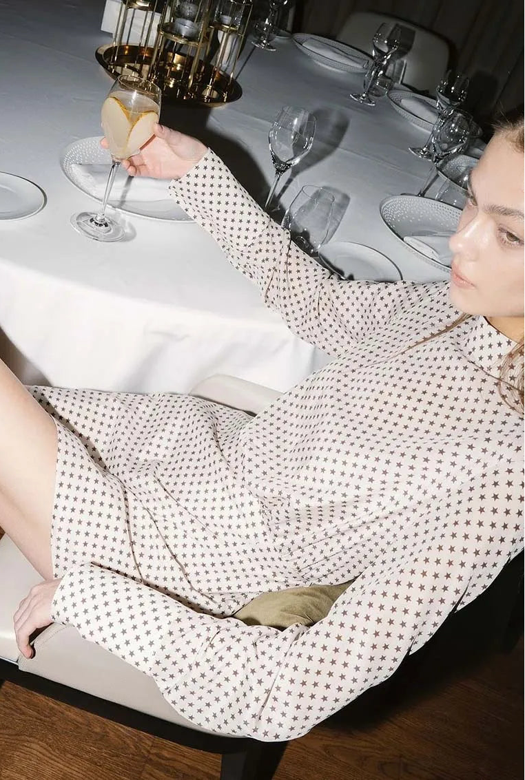 Woman sitting at a dining table with a glass of wine, wearing a polka dot dress.
