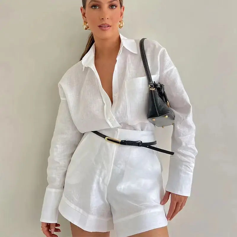 Woman wearing a white outfit with a black belt and handbag against a plain background