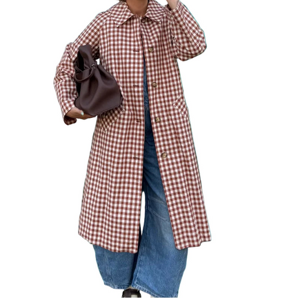 Woman wearing a checkered coat standing outdoors with plants and a building in the background