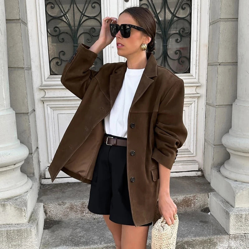 Woman in a brown coat and black skirt standing in front of a building entrance.