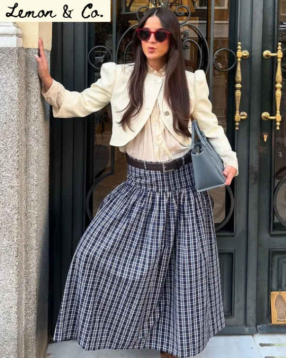 Woman in stylish outfit with plaid skirt and sunglasses standing outside a building.