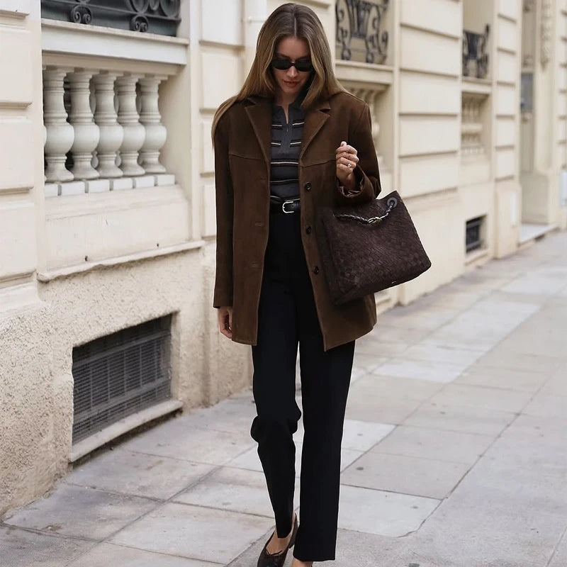 Woman in a brown coat and sunglasses holding a brown handbag on a city street.