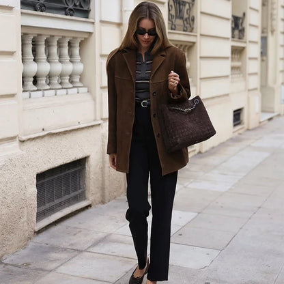 Woman in a brown coat and sunglasses holding a brown handbag on a city street.